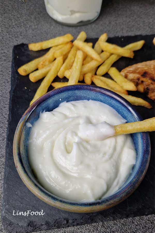 Close-up of creamy Lebanese toum in a blue bowl with a fry dipped inside, surrounded by more fries on a black slate board.