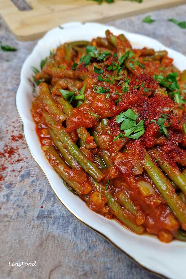 Close-up of loubieh bi zeit in an oval china platter, green beans coated in silky passata sauce, garnished with parsley and Aleppo flakes.