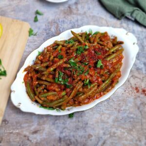Middle Eastern green beans in passata sauce (loubieh bi zeit) in an oval china bowl, with parsley and Aleppo flakes on top.