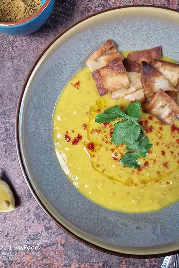 Close-up of shorbat adas in a green bowl, topped with cumin and chilli flakes, olive oil drizzle and lemon juice, with lemon wedges on the table.