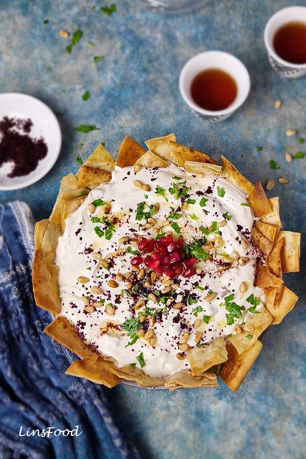 Overhead view of fattet hummus in a bowl lined with toasted pita and topped with yoghurt, pine nuts and parsley