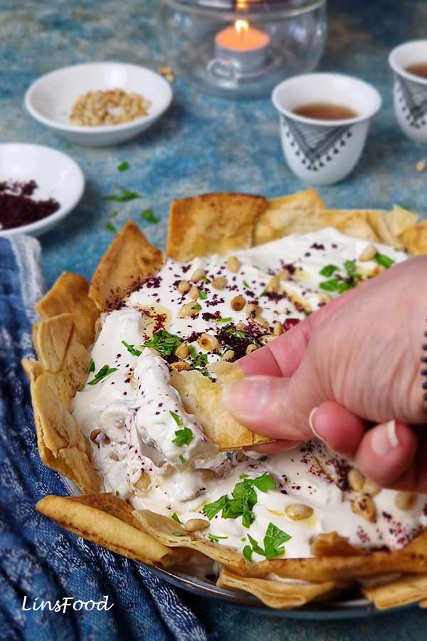 Hand dipping a piece of crispy pita into chickpea fatteh topped with yoghurt, parsley and pine nuts