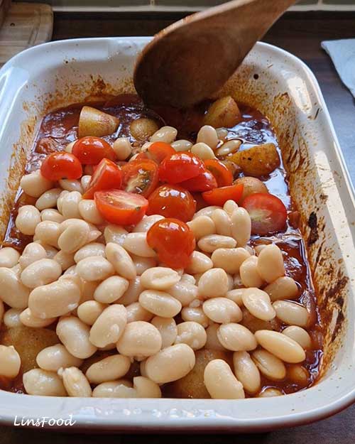 Butter beans and cherry tomatoes added to partially baked potatoes in tomato sauce for vegan fasolia traybake.