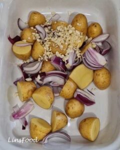 Potatoes, red onion wedges and chopped garlic in a white roasting dish for vegan fasolia traybake.