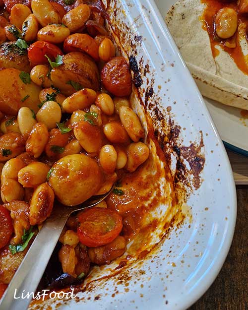 Spoon serving vegan fasolia traybake with butter beans, potatoes and tomato sauce beside flatbread.
