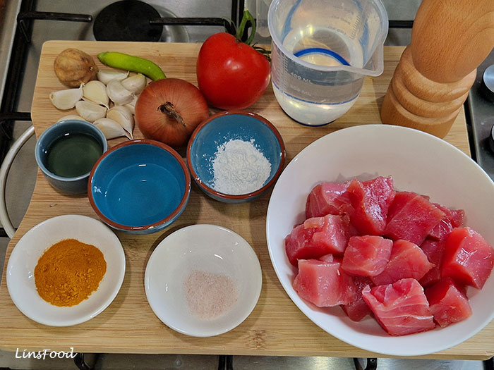 The ingredients for Omani paplou arranged on a chopping board, including fresh tuna chunks, tomatoes, onion, garlic, green chilli, brown dried lime, turmeric, salt, pepper, cornflour, lime juice (not in image) and water.