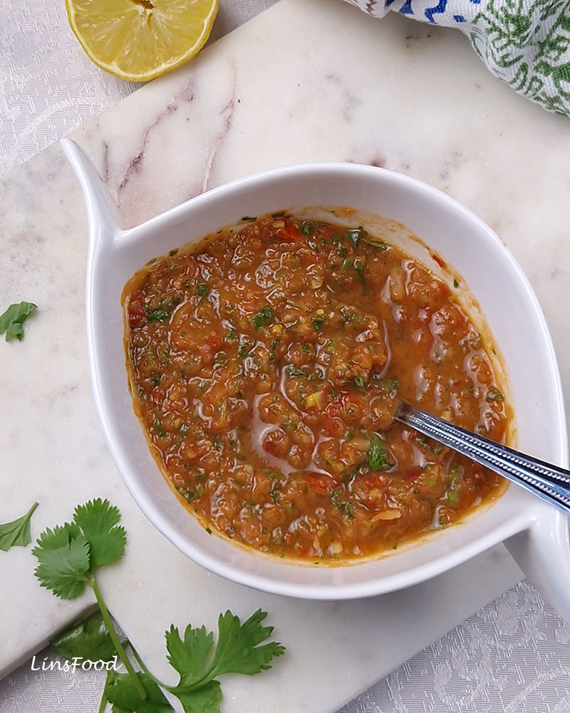 An overhead view of reddish-brown daqqus in a white bowl with a silver spoon, surrounded by coriander leaves, half a lemon and a patterned cloth.