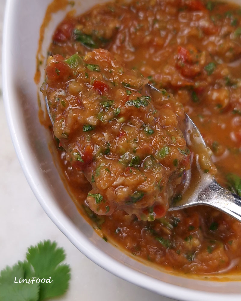 A silver spoon holding reddish-brown daqqus with visible pieces of tomato, chilli and chopped coriander, lifted above a white bowl of the salsa.