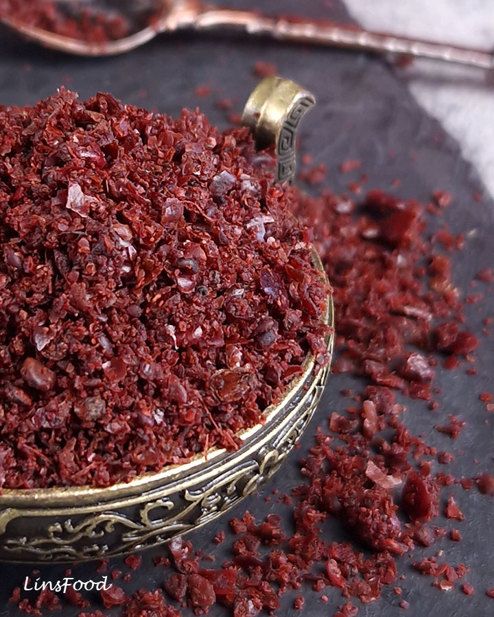 Extreme close-up of coarse Palestinian sumac flakes in an engraved brass bowl, with scattered pieces on a slate surface.