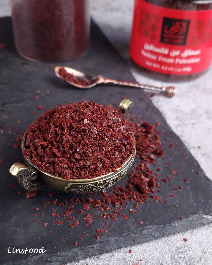 Coarsely ground Palestinian sumac in a decorative brass bowl on dark slate, with a jar of sumac in the background.